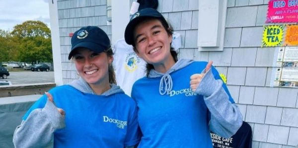 Two smiling women in blue Dockside Cafe shirts giving thumbs up outdoors.