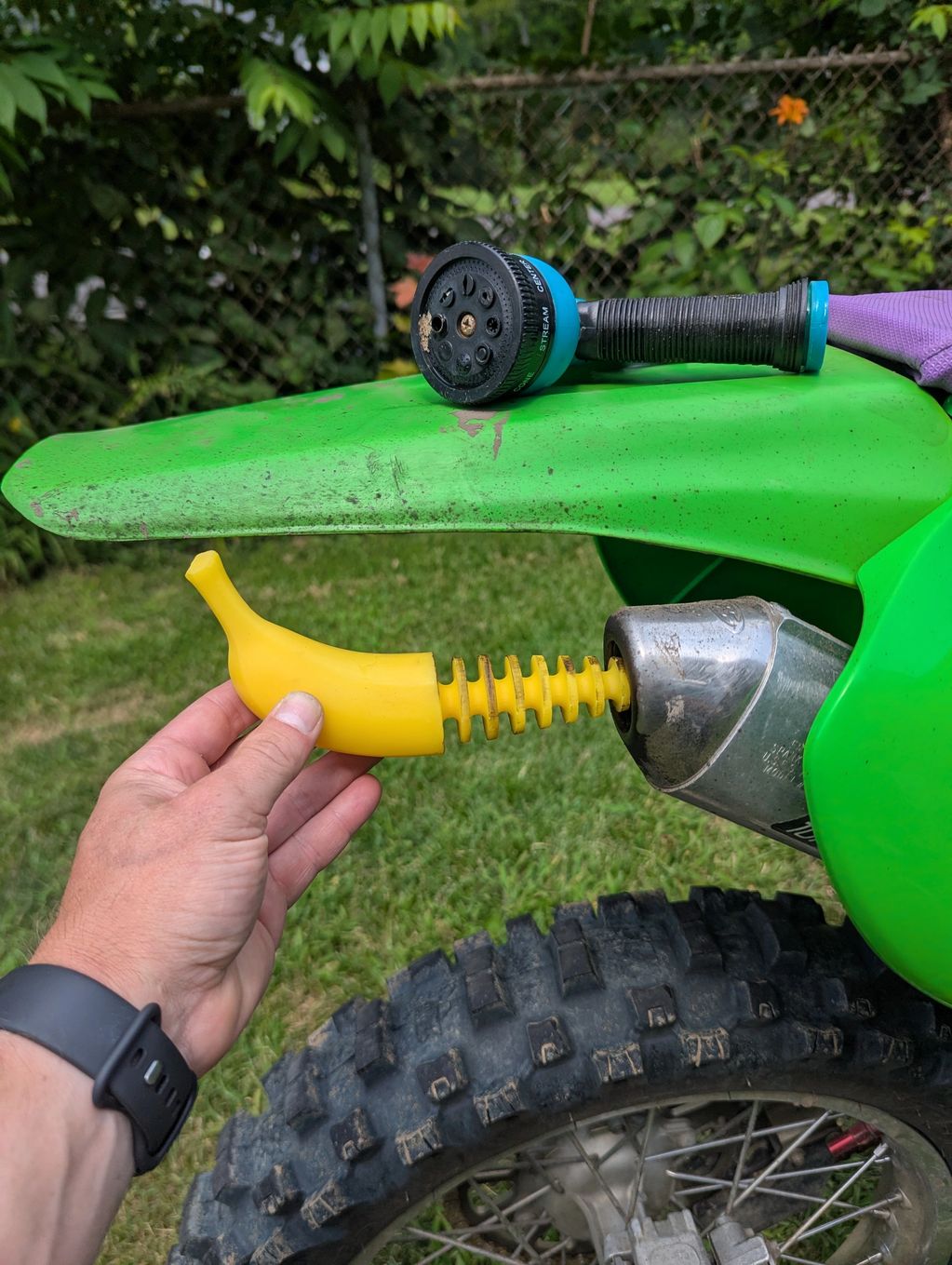 Hand holding a yellow banana-shaped exhaust cover near a green dirt bike exhaust.