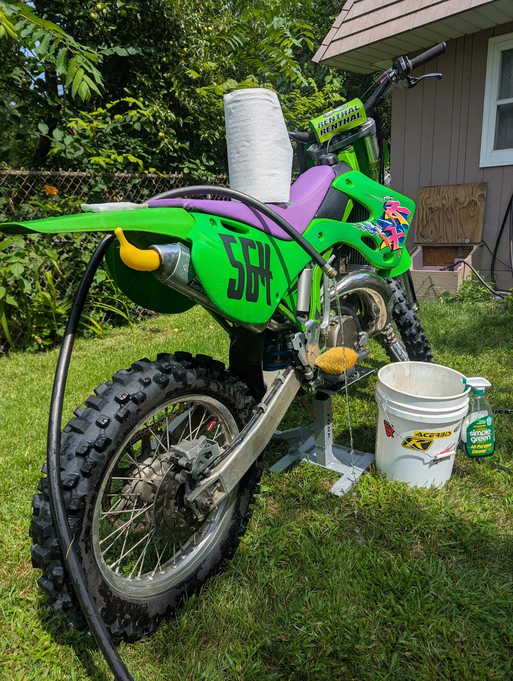 Green dirt bike being washed outdoors with paper towels, bucket, and cleaner.
