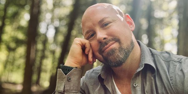 A man with a beard smiling thoughtfully in a sunlit forest.