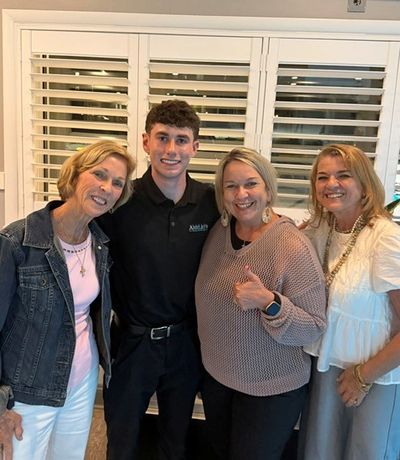Four people smiling together indoors in front of window shutters.