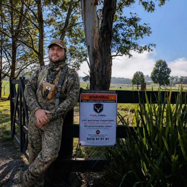 Man in camouflage leaning on a gate with a feral animal control warning sign.