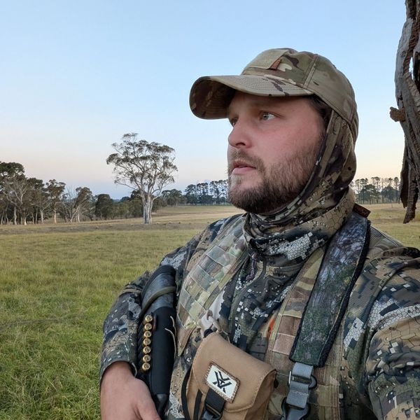 Man in camouflage gear holding a shotgun in a grassy field.