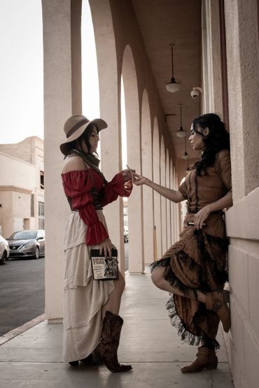 Two women in vintage Western attire exchange a cigarette outside a building with arches.
