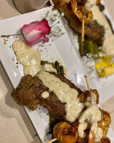 Steak topped with creamy sauce and shrimp skewers, garnished with a rose and herbs.