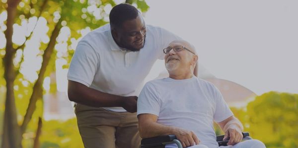 Caregiver smiling warmly at elderly man in wheelchair outdoors.