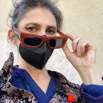 A person in a mask with hair blowing in the wind looks at the camera over brown sunglasses