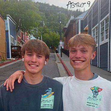 Two smiling teenage boys posing with arms around each other on a street with colorful buildings.