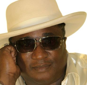 Man wearing a cream hat, sunglasses, and shiny cream shirt.