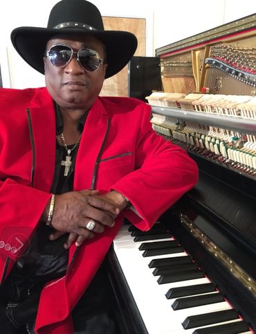 Man in red jacket and black hat posing by an open piano.