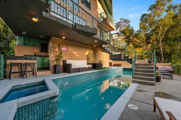 Modern backyard pool area with seating, bar, and lush greenery under a clear sky.