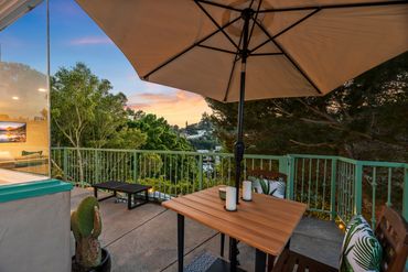 Cozy balcony setup with wooden table, umbrella, and sunset view over lush greenery.