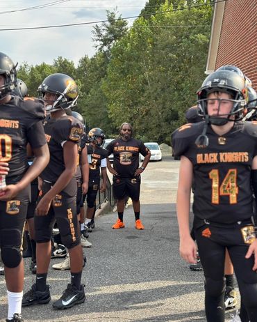 Youth football team Black Knights lined up with coach in bright orange shoes.