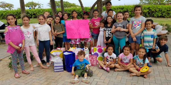 Group of children with gifts and a 'With Love 4 You!' sign outdoors.