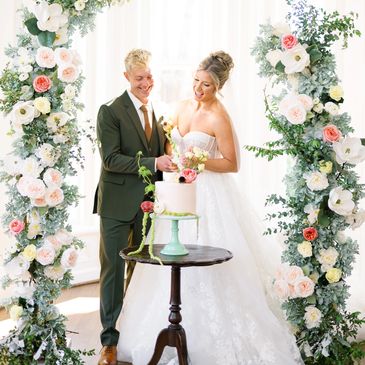 Bride and groom cutting wedding cake under floral arch.