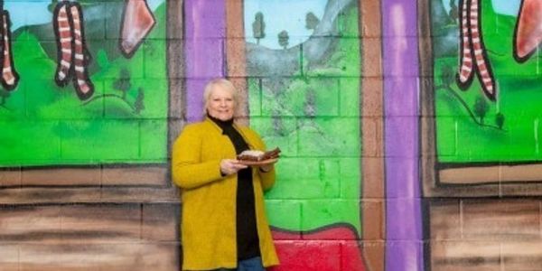 Woman in yellow coat holding food in front of colorful wall mural.