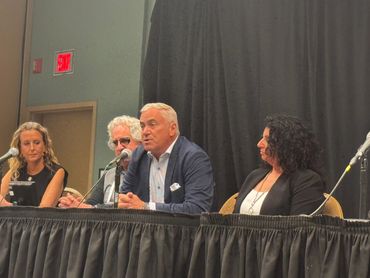 A speaker speaks to the microphone while other panelists listen in and watch him