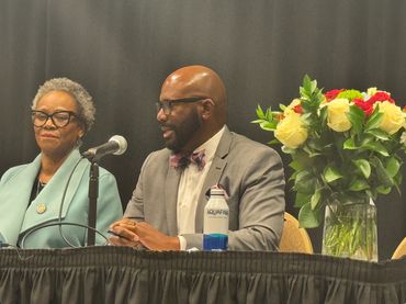 An Assemblymember in a bow tie speaking into a microphone while another listens in flowers on table
