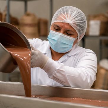 A worker pouring melted chocolate in a factory, wearing protective clothing.
