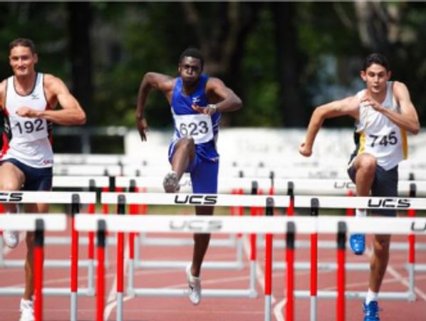 Three male athletes competing in a hurdles race on a track.