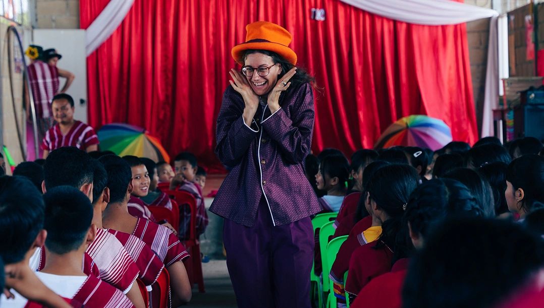 A woman in an orange hat joyfully engages with an audience in colorful attire.