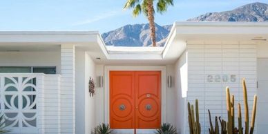 Modern white house with vibrant orange double doors and desert plants.