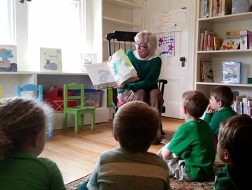 An elderly woman reads a picture book to a group of children in a cozy room.