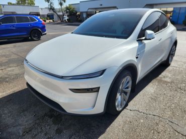 White Tesla Model Y parked in a lot with a blue SUV nearby.