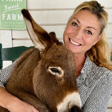 Woman smiling with a brown donkey on her lap.