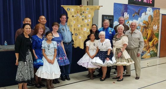 Group of people in traditional dresses posing with a decorated banner indoors.
