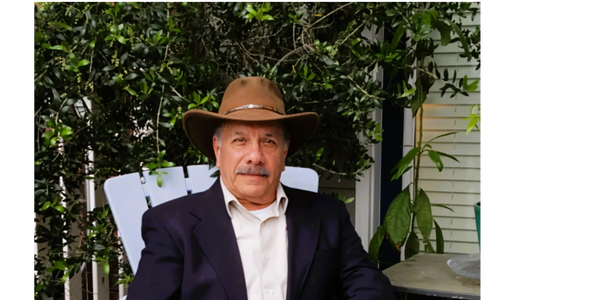 Man in a cowboy hat and blazer sitting outdoors on a white chair.