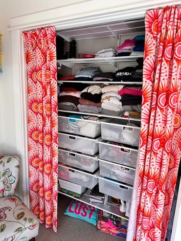 Organized closet with folded clothes and mesh drawers behind vibrant red-orange curtains.