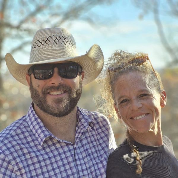 Smiling couple outdoors, man in cowboy hat and sunglasses, woman with curly hair and glasses on head.