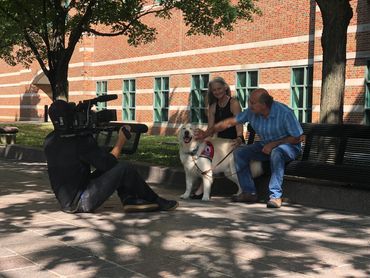 Two smiling people petting a fluffy white service dog are being filmed by a TV station