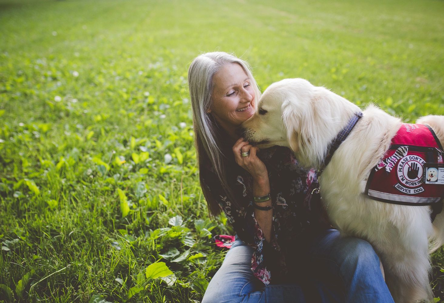A smiling woman is sitting in green grass with a fluffy white service dog 