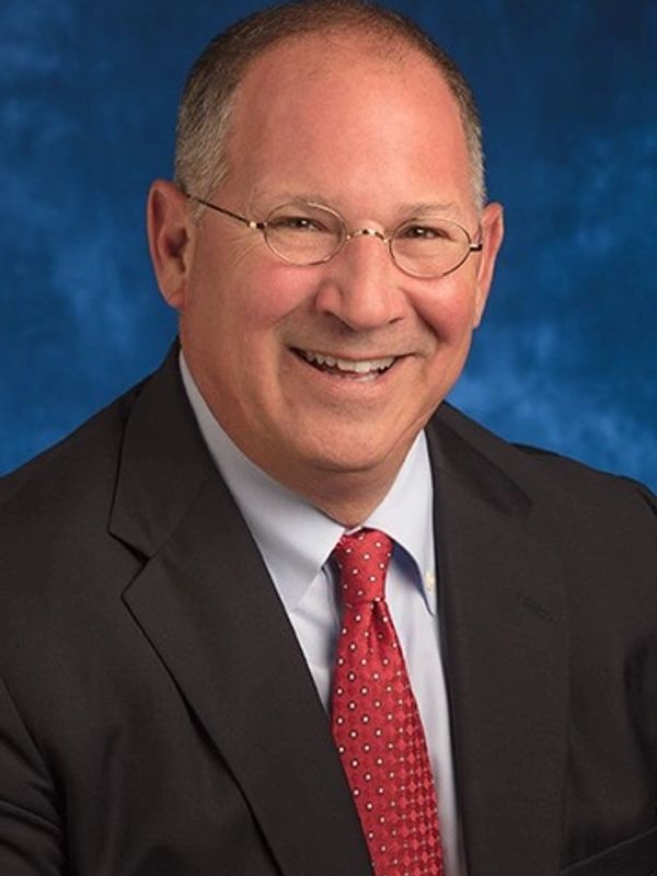 Smiling man in suit with glasses and red tie against blue backdrop.