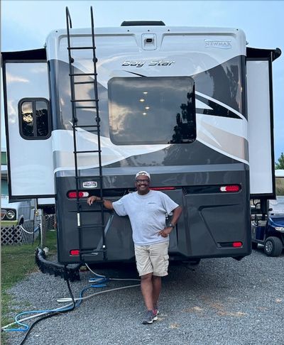 Man smiling and posing in front of a large RV with a ladder.