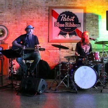 Two musicians perform on stage with guitar and drums under colorful lights.