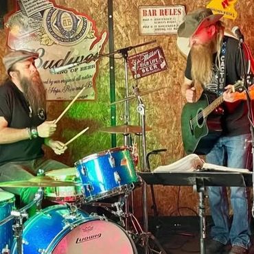 Two musicians with long beards play drums and guitar in a cozy bar setting.
