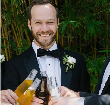 Groom in tuxedo toasting with friends outdoors.