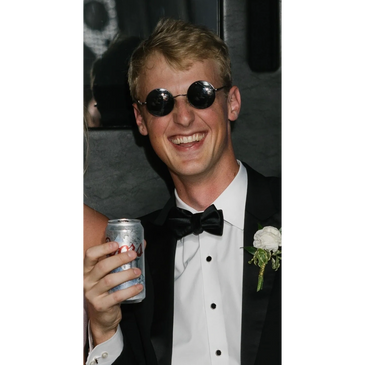 Young man in tuxedo and round sunglasses holds a can, smiling widely at the camera.