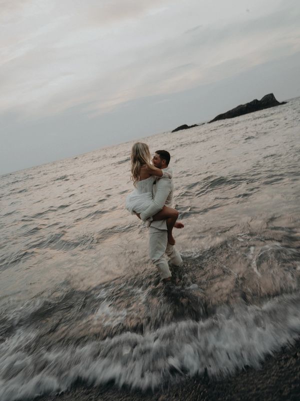 Man carrying woman in ocean water at sunset.