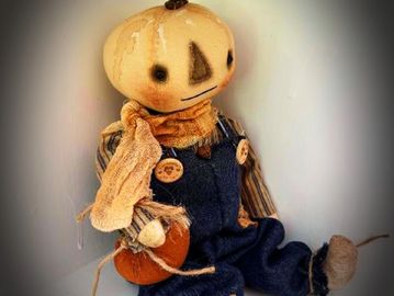A handmade scarecrow doll with a pumpkin head and denim overalls.