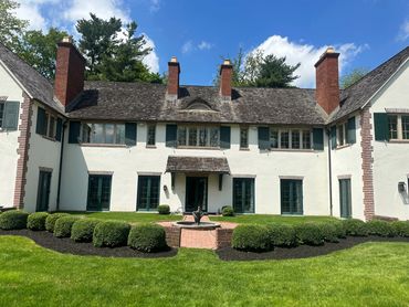 Large white house with green shutters and manicured bushes under a blue sky.