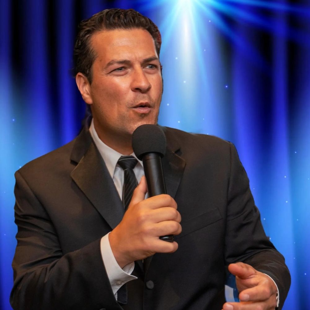 Lou Dottoli singing into a microphone with a blue stage curtain background.