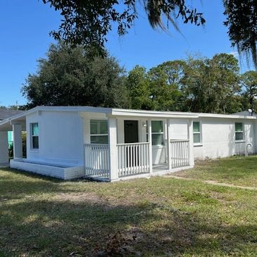 Single-story white house with a small front porch and grassy yard under clear blue sky.