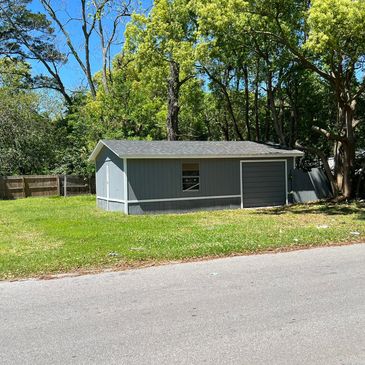 A small gray shed with a garage door on a grassy lot beside a road.