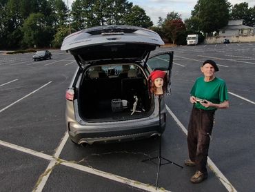 An elderly man stands near an open car trunk with a mannequin head on a stand.