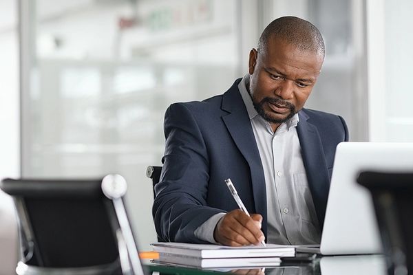 A focused businessman writing notes at his desk with a laptop.