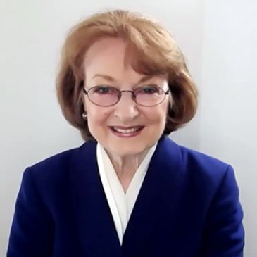 Smiling elderly woman in glasses and blue blazer with arms crossed.